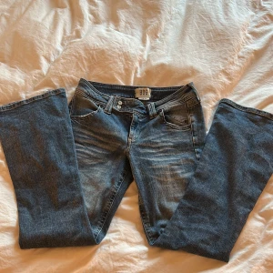 Blå bootcut jeans från BDG - Säljer ett par klassiska blå jeans från BDG med bootcut passform. Jeansen har låg midja, fem fickor och snygga slitningar framtill. Materialet är mjukt denim i bomull och de har dubbla knappar i midjan för extra detalj. MIDJEMÅTT 38 cm, innerbenslängd 76 