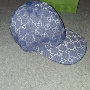 Blå kepsi med GG-mönster - Säljer en blå keps med ikoniskt GG-mönster i vitt över hela kepsen. Kepsen har böjd skärm, justerbart spänne bak i blått skinn och en liten guldfärgad GG-detalj på sidan. Materialet är bomull och skinn, vilket ger en lyxig känsla.
