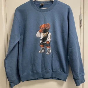 Blå sweatshirt med coolt björnmotiv - Blå sweatshirt med ett stort tryck av en björn i streetwear på framsidan. Tröjan har rund hals och långa ärmar, tillverkad i mjuk bomull. Perfekt för dig som gillar unika och roliga prints.