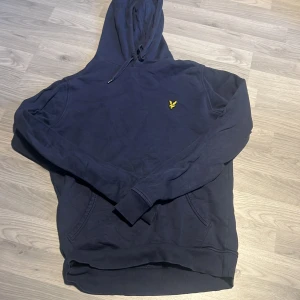 Mörkblå hoodie från Lyle & Scott - Mörkblå hoodie från Lyle & Scott med klassisk gul broderad logga på bröstet. Tröjan har huva med snörning, känguruficka och ribbade muddar vid ärmslut och nederkant. Perfekt för chill dagar eller när du vill ha en clean streetstyle.