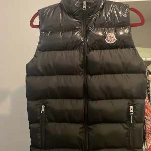 Svart dunväst från Moncler - Säljer en svart, glansig dunväst från Moncler med hög krage och dragkedja framtill. Västen har två fickor med dragkedja och klassisk Moncler-logga på bröstet. Perfekt för lager-på-lager och snygg till streetwear. Helt ny. Fick i present 😃