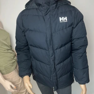 Svart pufferjacka från Helly Hansen - Säljer en svart pufferjacka från Helly Hansen med logga på bröstet och broderad text på kragen. Jackan har huva, dragkedja framtill och är vadderad för extra värme. Perfekt för kalla dagar och enkel att styla till det mesta.