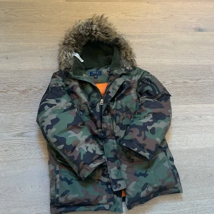 Polo Ralph Lauren Jacka - Parkajacka från Polo Ralph Lauren i camomönster med fuskpäls på huvan och grön fleecefoder. Jackan har flera fickor, dragkedja och knappar framtill samt patch på ärmen. Perfekt för kalla dagar och riktigt snygg streetstil.