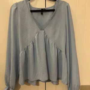 Ljusblå blus från Vero Moda - Supersöt ljusblå blus från Vero Moda med v-ringning och spetsdetalj vid halsen. Blusen har långa ärmar med resår vid ärmslut och volang nedtill som ger en flowy känsla.  