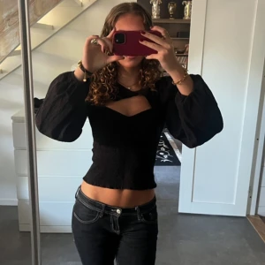 Svart topp med ballongärmar från Envii - Säljer en svart blus från Envii med smockad kropp och stora ballongärmar. Toppen har en snygg cut-out detalj vid bröstet. Den är ribbad vid magen och ryggen