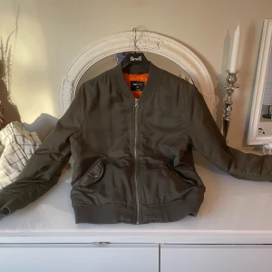 Olivgrön bomberjacka från FB Sister XL - Snygg olivgrön bomberjacka från FB Sister i storlek XL. Jackan har klassisk ribbad krage, muddar och nederkant, samt orange quiltat foder. Utrustad med dragkedja framtill och två fickor med knappstängning. Perfekt för dig som gillar streetstyle.
