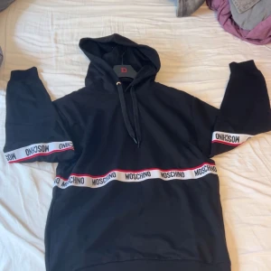 Svart Moschino hoodie med logoband - Inköpt för 2700, helt ny aldrig använd? Testade den och den var för stor. Svart hoodie från Moschino med huva och snörning. Tröjan har vita och röda logoband med Moschino-tryck runt bröstet och ärmarna. Klassisk passform och ribbade muddar. Perfekt för dig som gillar streetwear och vill sticka ut med en snygg logga.