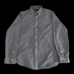 Ralph Lauren skjorta - Klassisk blå och vit rutig slim fit skjorta från Ralph Lauren med gul broderad logga på bröstet. Skjortan har lång ärm, knappar hela vägen och button down-krage. Perfekt för dig som gillar stilren och tidlös design.