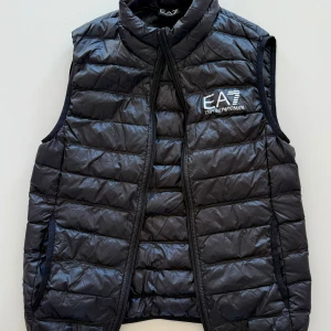 Svart dunväst från Emporio Armani - Snygg svart dunväst från Emporio Armani EA7 med quiltad design och dragkedja framtill. Västen har hög krage och logga broderad på bröstet. 