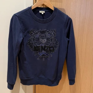 Mörkblå Kenzo sweatshirt med tiger - Säljer en mörkblå sweatshirt från Kenzo med ikonisk broderad tiger och pärldetaljer på bröstet. Tröjan har rund hals, ribbade muddar och är långärmad. Perfekt för dig som vill sticka ut med en cool och trendig look.