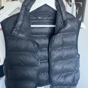 Svart dunväst från Zara - Säljer en svart croppad dunväst från Zara i storlek S. Västen är välanvänd och har synligare sminkfläckar på kragen samt en liten fläck vid ena fickan. Västen saknar även en snördetalj i kragen. Plagget har inga andra defekter eller skador på sig. Finns även så man kan reglera passformen inuti plagget. För referens är jag 164cm.