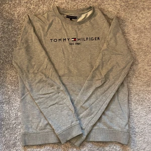 Grå sweatshirt från Tommy Hilfiger - Säljer en stilren grå sweatshirt från Tommy Hilfiger med broderad logga och text på bröstet. Tröjan har rund halsringning, långa ärmar och ribbade muddar. Perfekt för en avslappnad och snygg look.