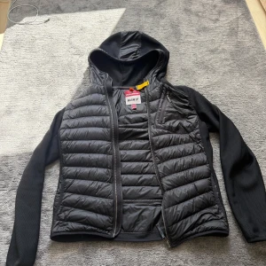 Svart pufferjacka från Parajumpers - Säljer en svart pufferjacka från Parajumpers med huva och snygga quiltade paneler. Jackan har dragkedja framtill, långärmade meshpartier och coola detaljer som bröstficka med zip och orange tag. Perfekt för dig som gillar street och funktion.