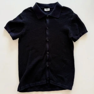 Svart kortärmad skjorta från Kappahl - Svart skjorta från Kappahl med korta ärmar och klassisk krage. Skjortan har en strukturerad yta och knäppning framtill med svarta knappar. Perfekt för en clean och stilren look.