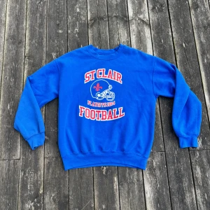 Blå St Clair Football sweatshirt - Blå sweatshirt med tryck 'St Clair Football Playoffs 2014' i rött och vitt framtill. Klassisk crewneck-modell med långa ärmar och ribbade muddar. Perfekt för dig som gillar sportig stil och college-vibe.