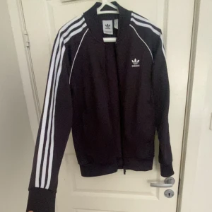 Adidas bomberjacka  - Fin vinbrun Adidas bomberjacka med vita ränder längs ärmarna och broderad logga på bröstet. Jackan har dragkedja framtill, ribbade muddar och är tillverkad i polyester. Perfekt för en sportig och stilren look. Pris kan diskuteras 