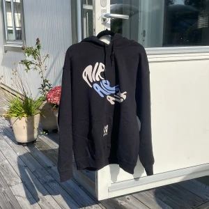 Svart hoodie Marcus & Martinus M - Svart hoodie från Marcus & Martinus med tryck i vitt och blått både fram och bak. Stor känguruficka och huva med snörning. Tillverkad i 50% bomull och 50% polyester. Perfekt för dig som gillar coola prints och streetstyle.