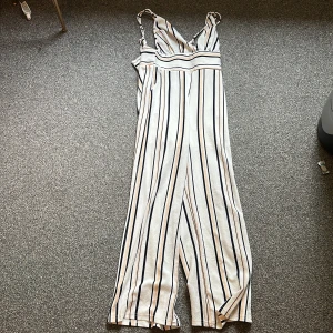 Vit och svart randig byxdress - Säljer en snygg byxdress med smala axelband och djup v-ringning. Den är vit med svarta och beige vertikala ränder, har markerad midja och vida ben. Perfekt för dig som gillar en trendig och luftig look. Materialet känns lätt och skönt mot huden.