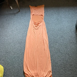 Ljus orange långklänning med cut-out - Säljer en snygg ljus orange långklänning med cut-out detalj över magen. Klänningen är ärmlös och har en kroppsnära passform som framhäver siluetten. Materialet är mjukt och stretchigt, perfekt för varma dagar. Enkel och stilren design som ger en cool vibe. Är 1.61 och är tyvärr för kort för denna klänning annars va den super fin! Passar för dig som är 1.67! 