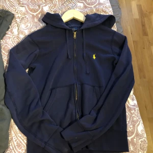 Marinblå hoodie från Polo Ralph Lauren - Säljer en marinblå hoodie från Polo Ralph Lauren med dragkedja och huva. Klassisk gul broderad logga på bröstet och fickor framtill. Skön bomullskvalitet och långärmad modell, perfekt för chill dagar.