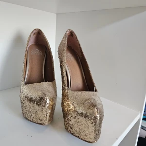 Glittriga guldiga pumps från Cuba Shoes - Säljer ett par riktigt snygga pumps i guld med glittrig yta från Cuba Shoes. Skorna har hög klack och plattform, rund tå och är perfekta för dig som vill sticka ut. Insidan är i beige/brunt syntetmaterial och utsidan är täckt av glittrande paljetter. 16cm