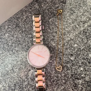 Michael Kors klocka i silver och guld - Stilren Michael Kors klocka med rund urtavla, silverfärgat armband med gulddetaljer och glittrande stenar runt boetten några stenar är borta men det kan man lösa på egen hand. Urtavlan är ljus med guldiga visare och index. Perfekt accessoar för dig som gillar en lyxig men clean look. Klockan är värd 4000kr. Ett Michael kors armband är också med i köpet. Pris kan diskuteras vid snabb köp. 