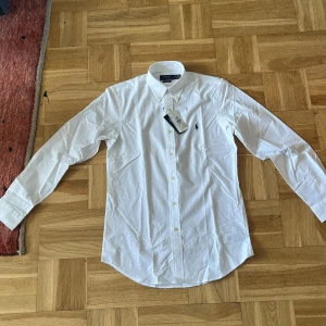 Vit skjorta från Polo Ralph Lauren S - Klassisk vit skjorta från Polo Ralph Lauren i bomull, custom fit. Skjortan har lång ärm, knappar framtill och den ikoniska broderade loggan i mörkblått på bröstet. Perfekt för en clean och stilren look.