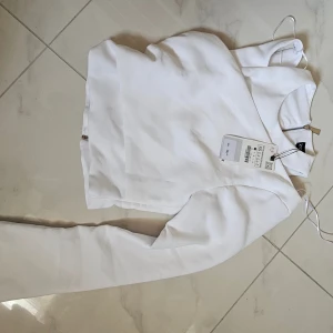 Vit långärmad topp från Zara XS - Säljer en stilren vit långärmad topp från Zara i storlek XS. Toppen har en rak passform, hög hals och dragkedja i ryggen som ger en clean look. Materialet är mjukt och lätt strukturerat, perfekt för att lyfta din outfit.