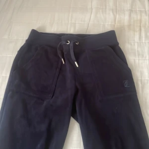 Mörkblå mjukisbyxor Juicy Couture XXS  - Snygga mörkblå mjukisbyxor från Juicy Couture i supermjuk velour. Byxorna har bred resår i midjan med snörning, stora fickor fram och bak med silverfärgade knappar. Perfekt för chill eller streetstyle. Diskret Juicy Couture-logo på ena fickan.