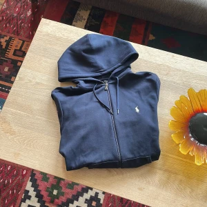 Mörkblå hoodie från Polo Ralph Lauren - Stilren mörkblå kofta från Polo Ralph Lauren med logga på bröstet. Perfekt nu till hösten. Köpt för 1600kr på room 1006, den är nopprig lite här och var men det går lätt att ta bort. Använts varsamt och har inga hål eller märkbara defekter.