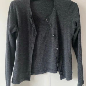 Mörkgrå kofta med knappar - En stilren mörkgrå kofta med lång ärm och knäppning framtill. Koftan är i ett mjukt, stickat material och har en klassisk rak passform. Perfekt att slänga över en t-shirt eller linne för en enkel och snygg look. Liten skjorta detalj nere vid ärmarna.