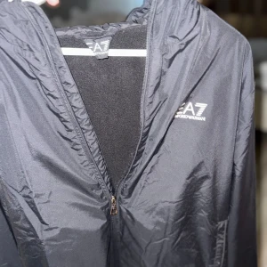 Svart vindjacka från EA7 Emporio Armani - Svart vindjacka från EA7 Emporio Armani med huva och dragkedja framtill. Jackan har en stilren design med EA7-logga på bröstet och elastiska muddar. Tillverkad i lätt polyester som passar perfekt för blåsiga dagar.