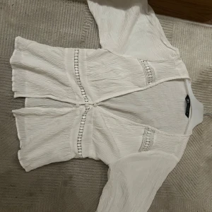 Vit blus med spetsdetaljer från Zara - Säljer en vit blus från Zara med fina spetsdetaljer och dekorativa knappar framtill. Blusen har lång ärm och är tillverkad i ett lätt, krinklat tyg som ger en avslappnad känsla. Perfekt att styla med jeans eller kjol för en fräsch look.