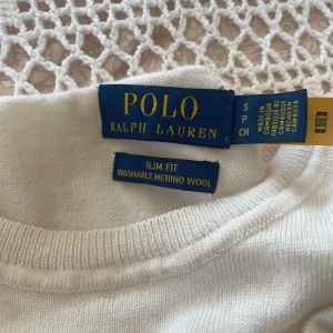 Vit merinoulltröja från Polo Ralph Lauren - Slim fit vit stickad tröja från Polo Ralph Lauren i mjuk och tvättbar merinoull. Klassisk rund hals och långärmad design med diskret broderad logga på bröstet. Perfekt för dig som gillar stilrena och tidlösa plagg.