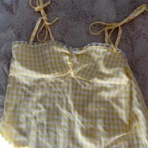 Gul och vit rutig sommarklänning - Supersöt gulvit rutig linnetopp med tunna knytband och spetsdetaljer längs kanten. Toppen har en liten rosett framtill och är gjord i ett lätt material som känns perfekt för varma dagar. Passar dig som gillar en romantisk och somrig stil.🫰🏼Kom inte till någon användning därför säljer jag den😉