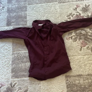 Authentic Vinröd skjorta från Yves Saint Laurent - Vinröd skjorta från Yves Saint Laurent i storlek L. Skjortan har lång ärm, klassisk krage och broderad YSL-logga med franska flaggans färger på ärmen. Tillverkad i mjuk bomull och har knappar framtill. Den sitter som M men är L, Pris kan diskuteras 