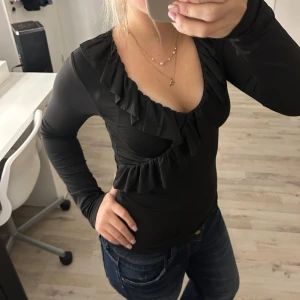 Svart volangblus från New Yorker  - Säljer en svart långärmad blus från Amisu med djup v-ringning och snygga volangdetaljer längs halsringningen och över bysten. Toppens material är stretchigt och följsamt, perfekt för dig som gillar en feminin och trendig look.