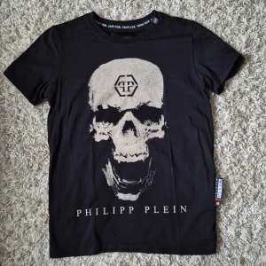 Svart t-shirt döskalle Philipp Plein Nyskick - Svart t-shirt med stor glittrande dödskalle framtill och logga både fram och bak. Klassisk passform med korta ärmar och rund hals. Tillverkad i mjuk bomull som känns skön mot huden. Snygg detalj med etikett i sidan. S/M