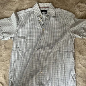 Rutig kortärmad skjorta från Eton - Kortärmad skjorta från Eton i vitt och ljusblått rutigt mönster. Skjortan har klassisk krage, knappar framtill och contemporary fit. Tillverkad i bomull för en skön och luftig känsla. Perfekt för dig som gillar stilren och clean look.