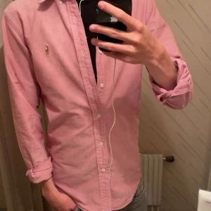 Rosa skjorta från Ralph Lauren - Säljer en klassisk rosa skjorta från Ralph Lauren med broderad logga på bröstet. Skjortan har lång ärm, knappar framtill och är gjord i mjuk bomull. Perfekt för dig som vill ha en stilren och tidlös look. Den är sydd innuti men syns inte när man har på sig den. Därav priset