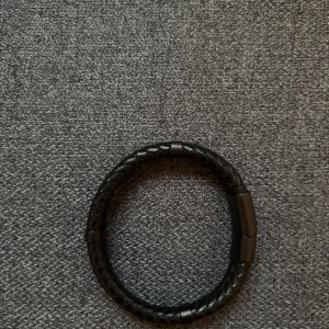 Svart flätat armband från USPA Ralph Lauren - Snyggt svart armband från USPA med flätad design och matt svart spänne. Armbandet har en stilren look och är tillverkat i konstläder med USPA-logga på spännet. Perfekt accessoar för att lyfta din outfit.