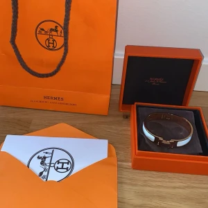 H vit armband med guldton - Exklusivt armband från H i klassisk design med vit emalj och guldfärgad metall. Armbandet har den ikoniska H-detaljen framtill och kommer i originalask med tillhörande påse och certifikat. Perfekt accessoar för dig som gillar lyxiga detaljer och stilren look.