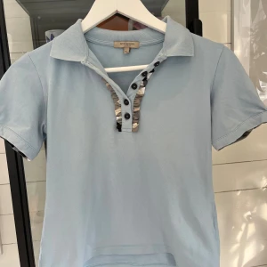 Ljusblå pikéblus från Burberry - Supersöt ljusblå pikéblus från Burberry med korta ärmar och puffdetalj med knapp. Krage och knäppning framtill med volangkant i grå, vit och svart. Perfekt för dig som gillar klassisk stil med en twist.