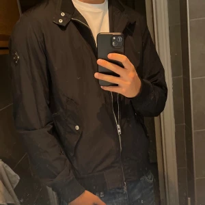 Svart vindjacka från J.Lindeberg - Svart vindjacka från J.Lindeberg med hög krage, dragkedja och snygga detaljer som logotyp på ärmen. Jackan har ribbade muddar och praktiska fickor framtill. Perfekt för dig som vill ha en stilren och sportig look, Nypris 2000 kr. 