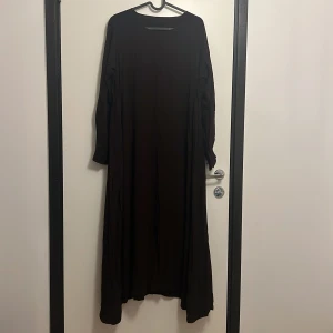 Mörkbrun långklänning med prickar - Långklänning i mörkbrunt med diskret prickigt mönster och lång ärm. Klänningen har rund halsringning och ett lätt, flowy tyg som ger en avslappnad vibe. Perfekt för dig som gillar en enkel men ändå unik look.