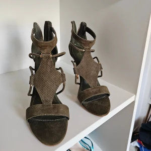 Olivgröna mockasandaler klackar från pimkie - Snygga olivgröna sandaler i mocka med höga klackar och öppen tå. Skorna har coola remmar med gulddetaljer, perforerat parti framtill och dragkedja bak för enkel påtagning. Perfekta för dig som vill sticka ut med en trendig och elegant look.