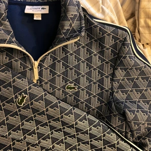 Blå mönstrad track suite från Lacoste - Säljer en blå och beige  track suiten från Lacoste med grafiskt mönster och klassisk krokodillogga på bröstet. Storlek S på tröja och M på byxor säljer pga för små. Knappt använda och köpte på NK i Göteborg. Skriv om det finns några funderingar🐊😁