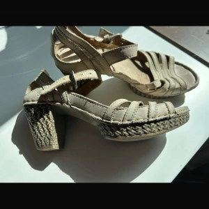 Beige sandaler med klack och flätad sula - Snygga beige sandaler med öppen tå och bred klack. Skorna har flera remmar i skinn och en justerbar rem med spänne runt ankeln. Klacken och sulan är dekorerade med flätad detalj som ger en boho-vibe. Perfekta för sommardagar när du vill ha både stil och komfort.