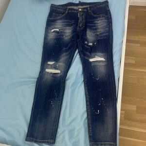 Dsquared2 blå distressed jeans - Snygga blå jeans från Dsquared2 med slitna detaljer, färgstänk i orange, blått och vitt samt klassisk femficksdesign. Jeansen har raka ben och tydliga slitningar framtill för en cool, trendig look. Perfekta för dig som vill sticka ut med din stil. Den är helt ny men morsan råka ta bort tagsen