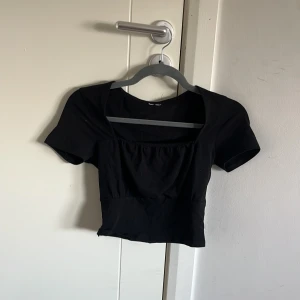 Svart croppad topp med fyrkantig ringning - Snygg svart croppad topp med korta ärmar och fyrkantig ringning. Toppen har en lätt rynkad detalj framtill och är figurnära i modellen. Perfekt att matcha med höga jeans eller kjol för en trendig look.